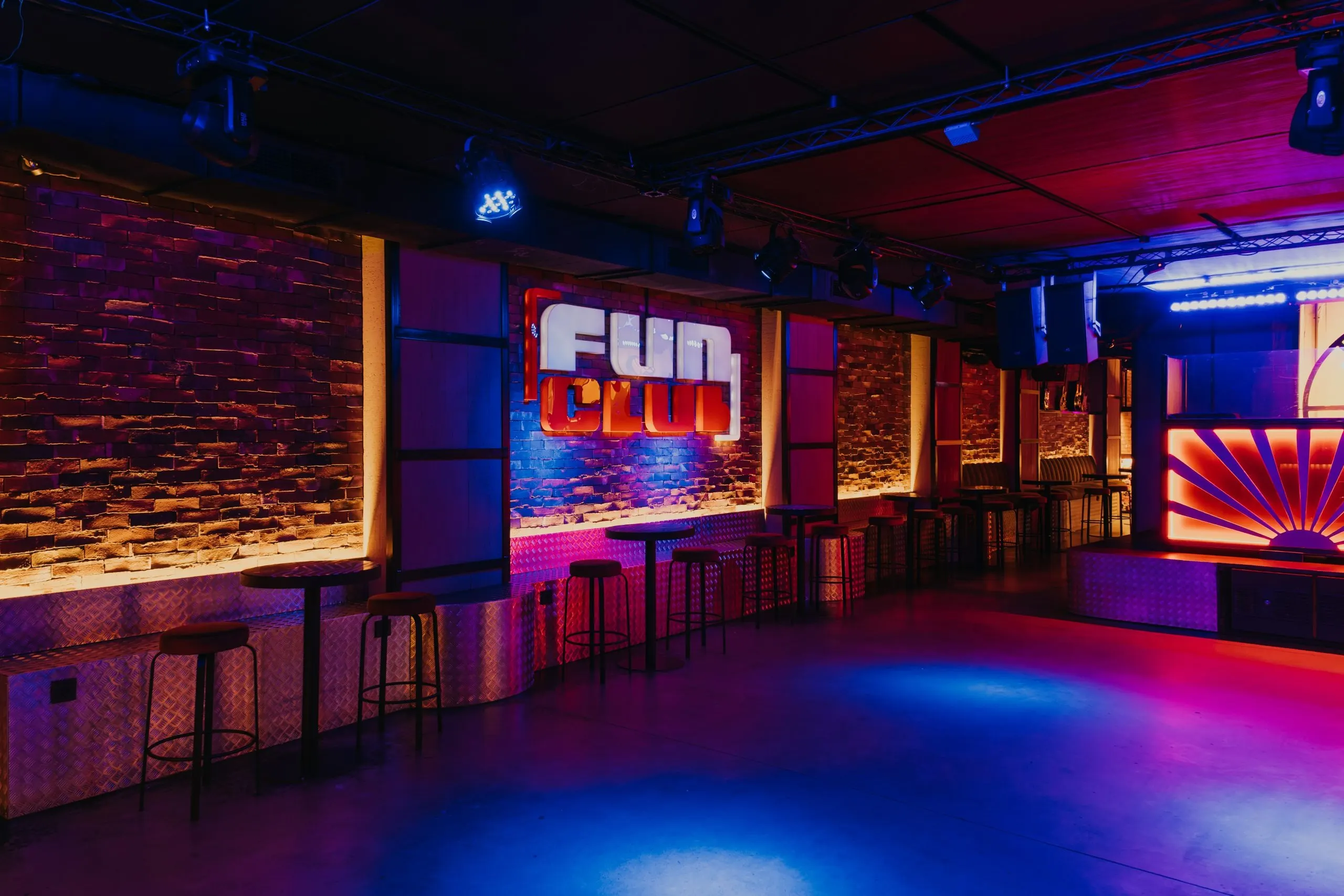 La imagen muestra un gran texto blanco “FUN CLUB” montado en una pared de ladrillos rojos. El texto está iluminado, proyectando un resplandor que se refleja en la pared y el área circundante.