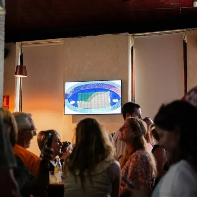 Pantalla con imagen de un estadio encendida en una sala llena de personas de pie, conversando y mirando en distintas direcciones.