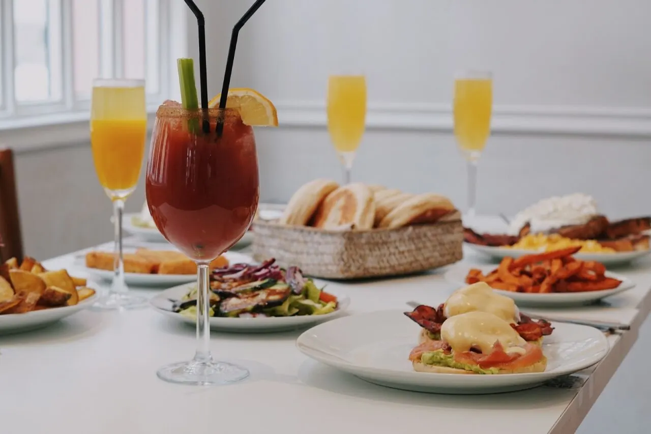 Mesa blanca con platos de brunch variados, cócteles Bloody Mary y mimosas, tostadas con huevos benedict y aguacate, batatas fritas y pan en cesta.