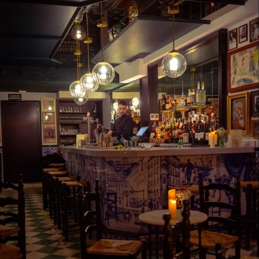 Interior de bar con suelo ajedrezado, mesas y sillas de madera. Barra decorada con arte, estanterías con botellas y lámparas esféricas colgantes.