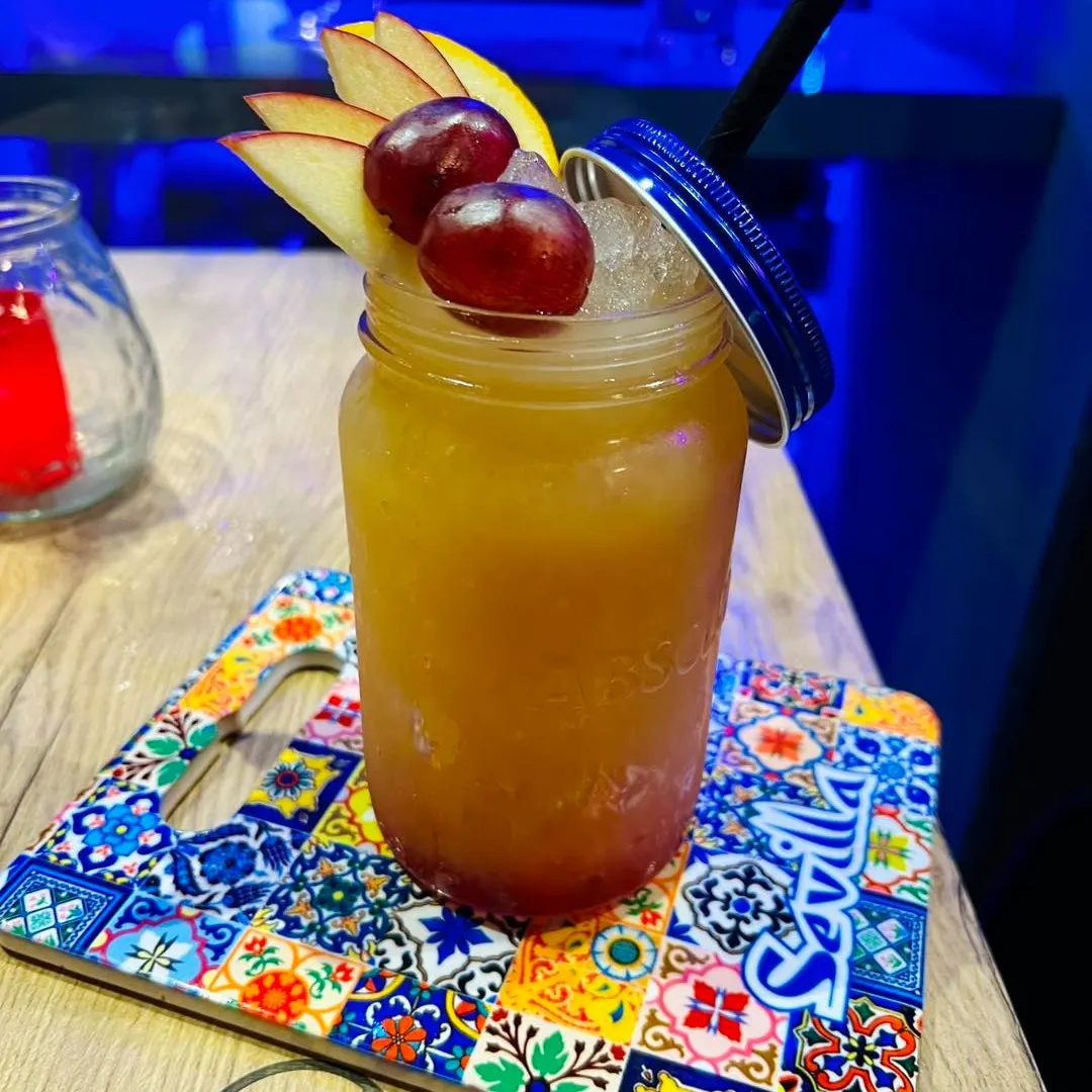 Un cóctel vibrante servido en un tarro, decorado con rodajas de manzana y uvas, sobre una colorida bandeja con motivos de Sevilla.
