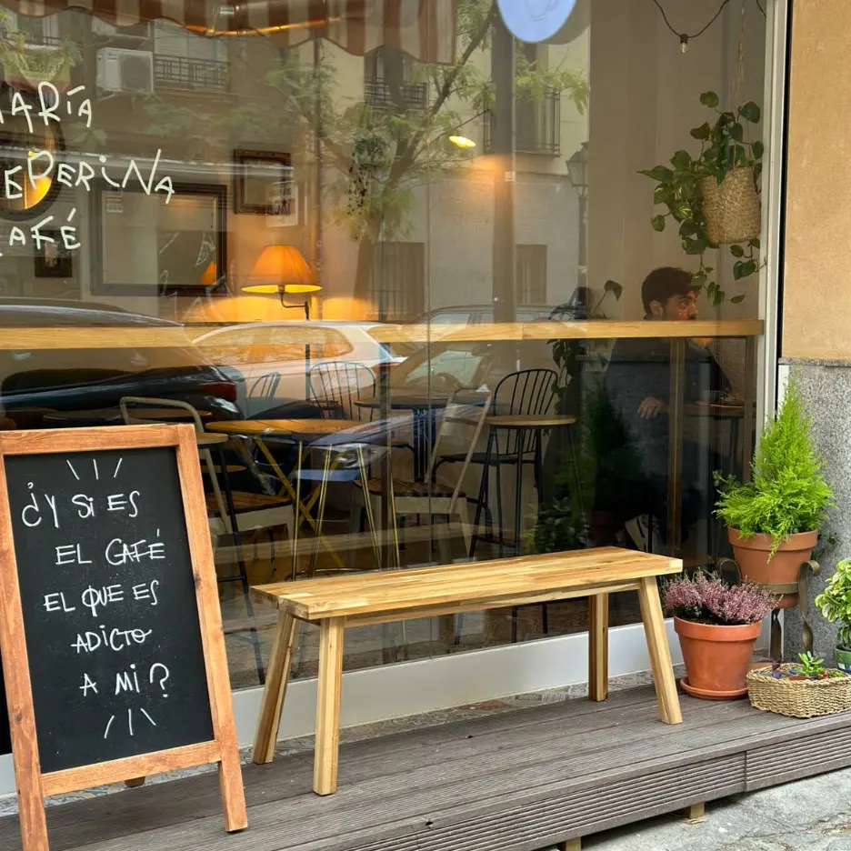 Exterior de cafetería con banco de madera y plantas en macetas. Cartel de pizarra con frase: “¿Y si es el café el que es adicto a mí?”.
