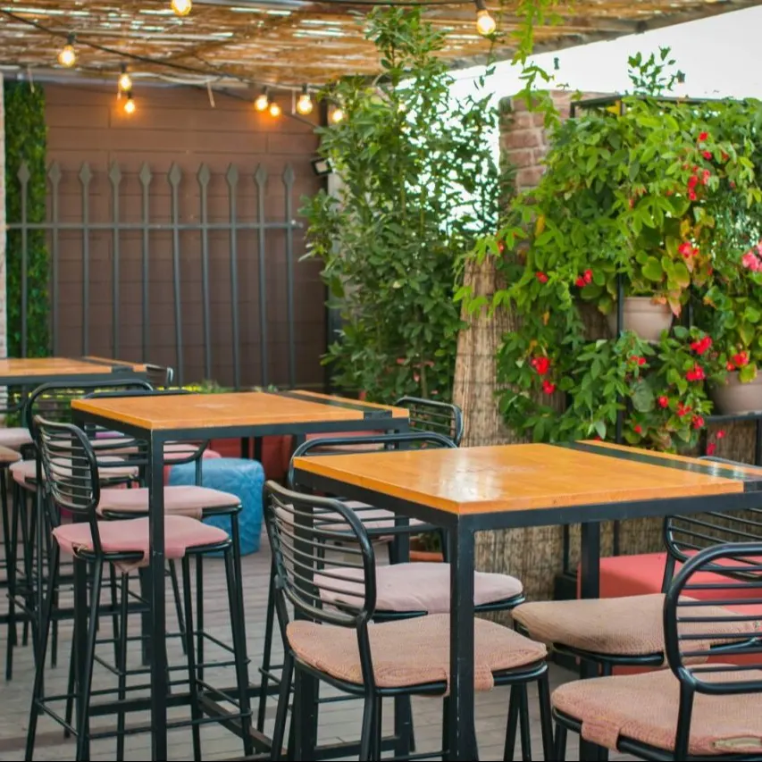 Zona exterior con mesas de madera y sillas negras acolchadas. Luces colgantes bajo pérgola, plantas verdes y flores rojas junto a muro de ladrillo.