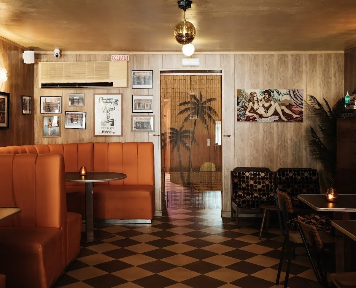 Cafetería retro con asientos naranjas, suelo ajedrezado, paredes con cuadros y cartel “ON AIR”. Decoración colorida y ambiente nostálgico.