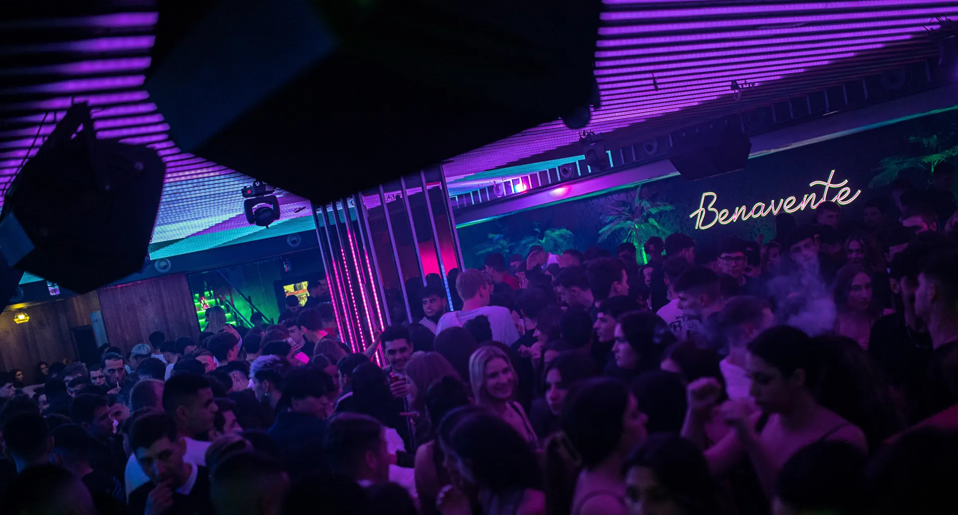 Ambiente nocturno en Benavente con iluminación violeta, multitud disfrutando y decoración moderna con estilo tropical.