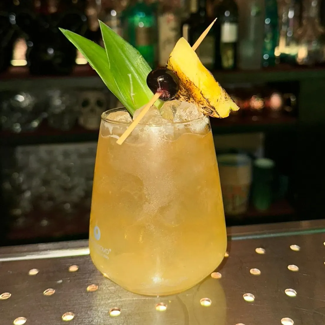 Copa de cristal con cóctel, hielo, menta y piña flambeada, sobre mesa de metal con estanterías con botellas de fondo.