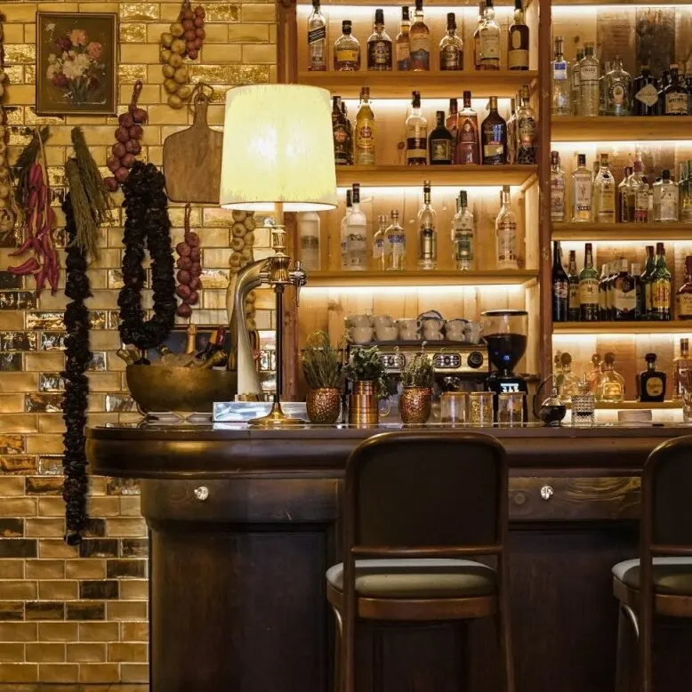Bar elegante con barra de madera, botellas iluminadas, taburetes altos y pared decorada con guindillas y ajos colgantes.