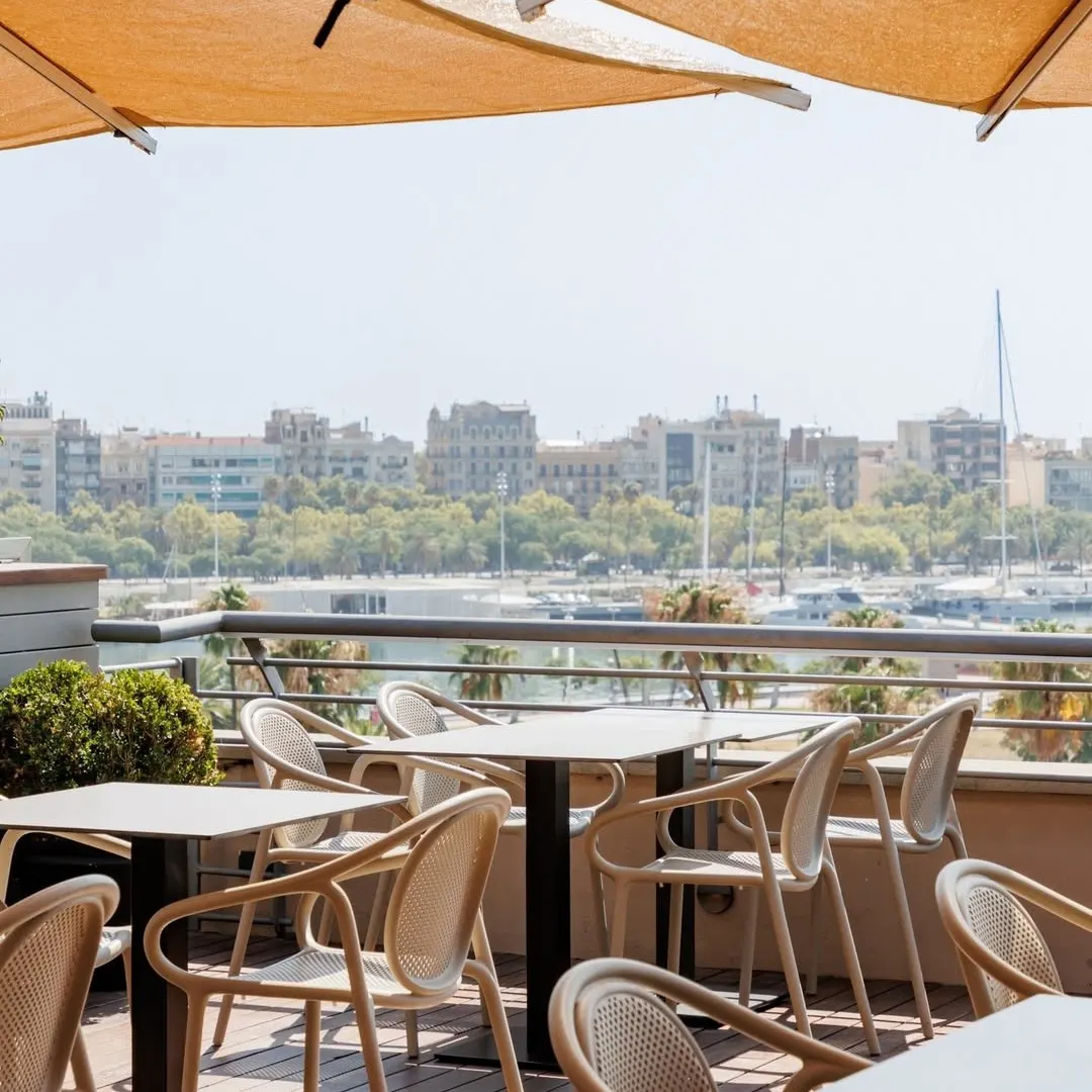 Terraza de La Duquesa de Cardona con vistas al puerto de Barcelona, mesas elegantes bajo sombrillas y una panorámica de la ciudad al fondo.