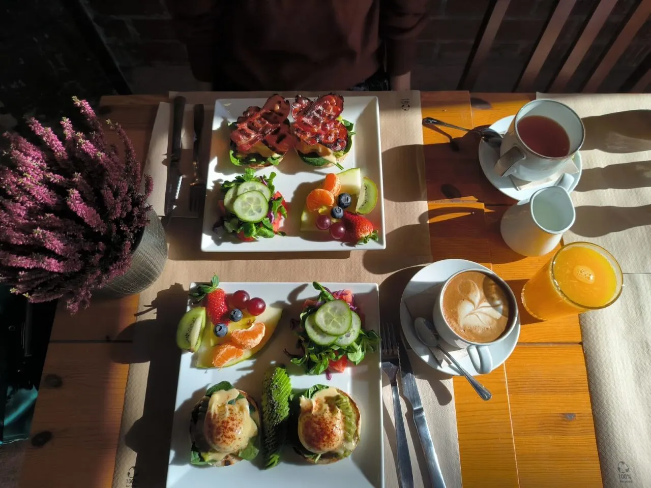 En la imagen se ve una mesa de madera con un brunch en Nolita, que incluye huevos benedictinos, tostadas con bacon, ensaladas con frutas, café, té y zumo de naranja.