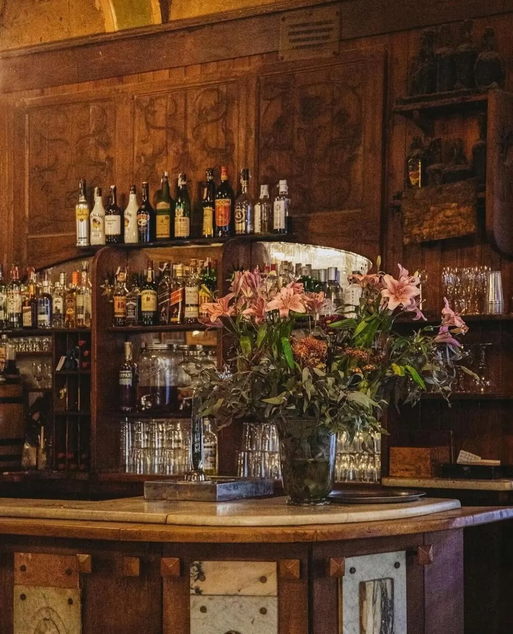 Bar rústico con mostrador de madera, estanterías llenas de botellas, ramo de flores en jarrón y ambiente cálido con decoración tradicional.