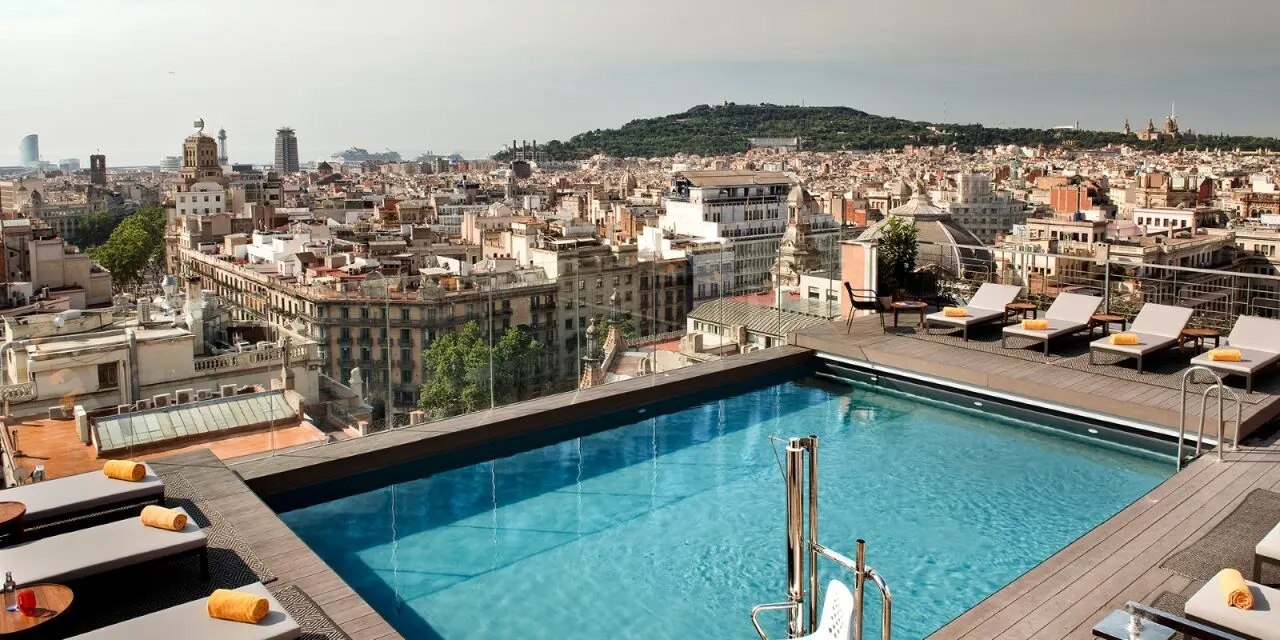 Piscina en azotea con tumbonas y toallas, vista panorámica de la ciudad con edificios y una colina al fondo. Ambiente relajado y urbano.