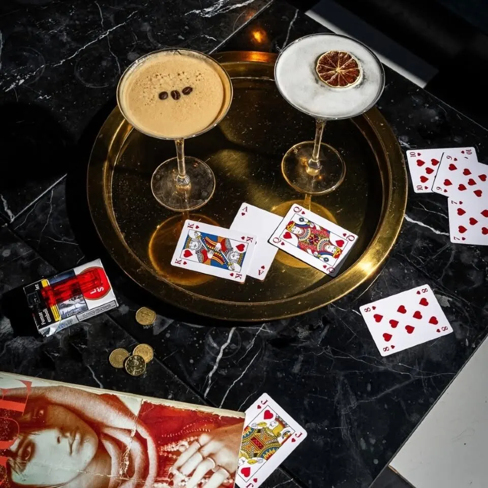 Mesa de mármol negro con cócteles, cartas, monedas, cigarrillos y revista; copa con café y otra con cítrico seco.