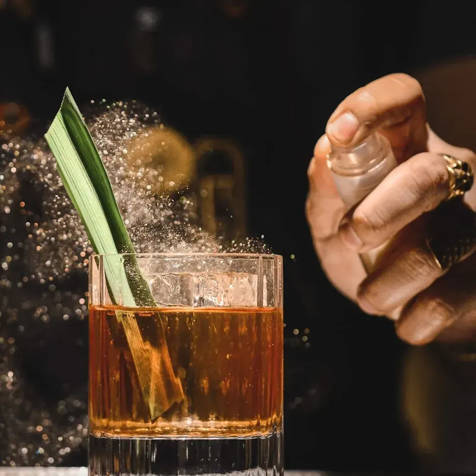 Cóctel en vaso bajo con hoja decorativa en Especiarium, mientras se rocía con un mist aromático que brilla bajo la luz.