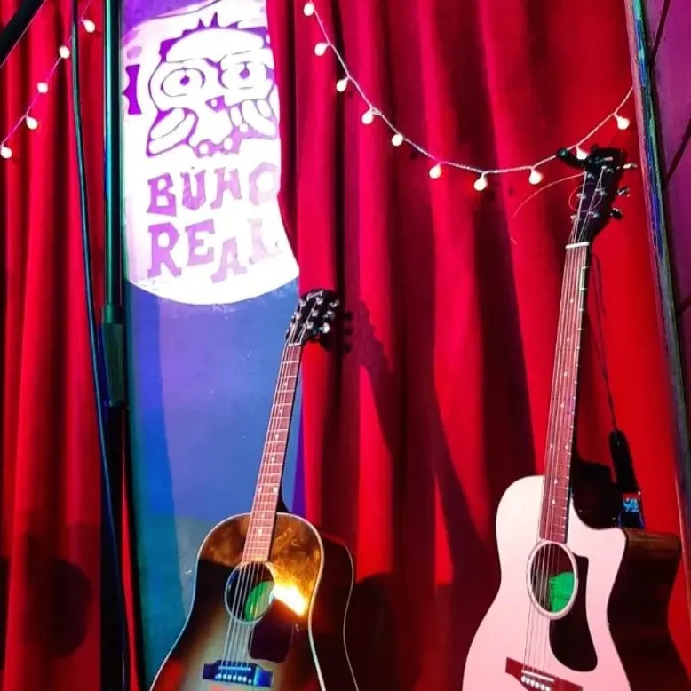 La foto muestra un escenario preparado para una actuación musical con dos guitarras acústicas colocadas en soportes, un micrófono en el centro, un taburete a la izquierda. El fondo presenta una pared de ladrillo rojo adornada con luces de cuerda.