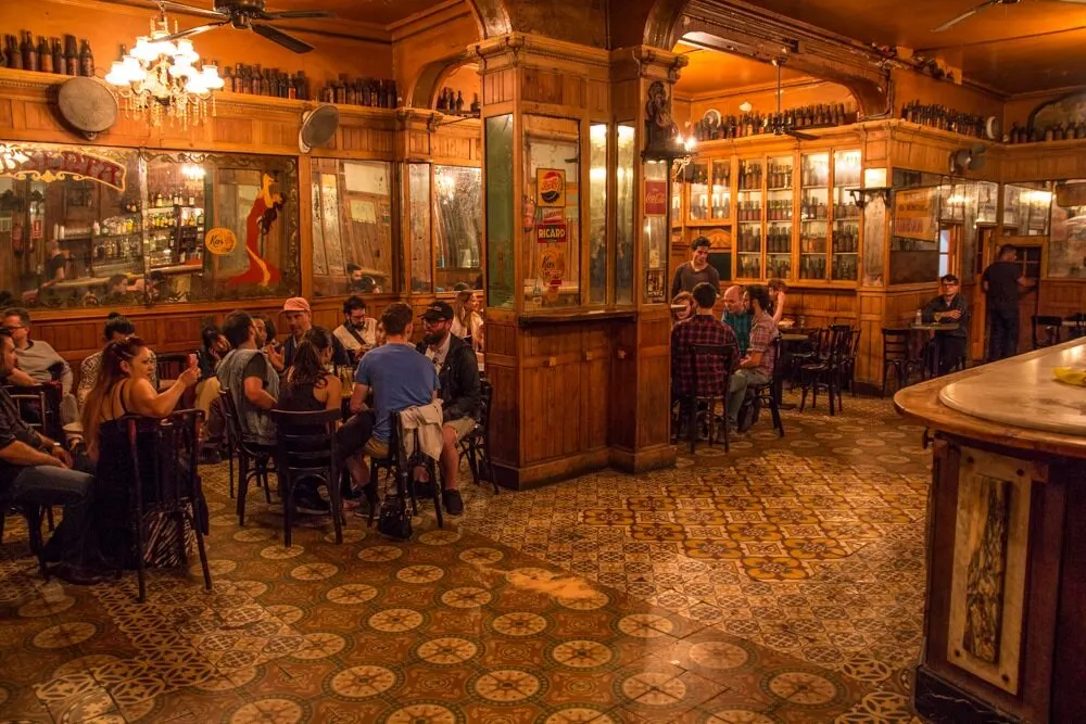 Bar vintage con muebles de madera, suelo de baldosas decorativas y estanterías con botellas. Gente conversando en un ambiente cálido y acogedor.
