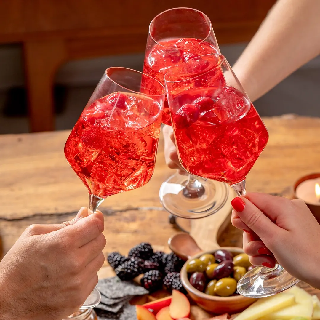 Brindis con tres copas de cóctel rojo sobre un tablero de madera lleno de quesos, embutidos, frutas y frutos secos, en un ambiente cálido de reunión