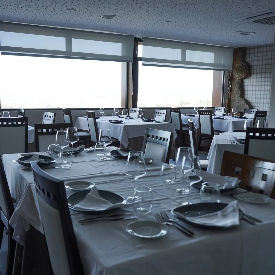 Salón del restaurante con manteles blancos, vajilla elegante, copas de vino y grandes ventanales con vistas al fondo, decorado con piedra y madera.