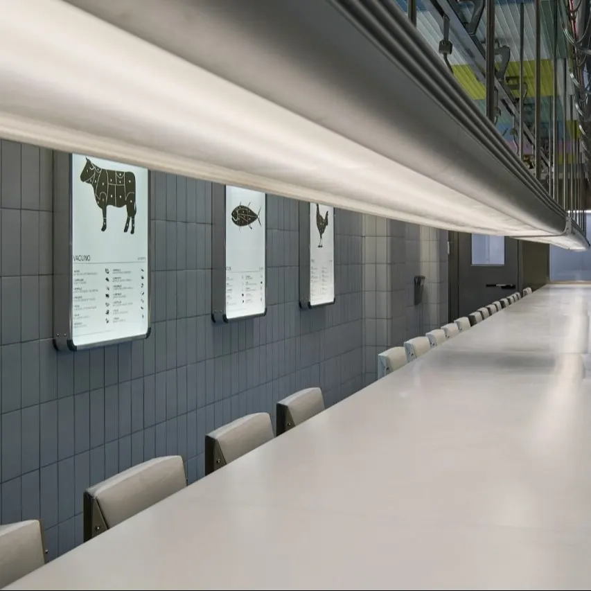 Mesa larga y moderna con sillas a ambos lados. Paredes con azulejos y carteles iluminados que muestran cortes de carne de vaca, pollo y pescado.