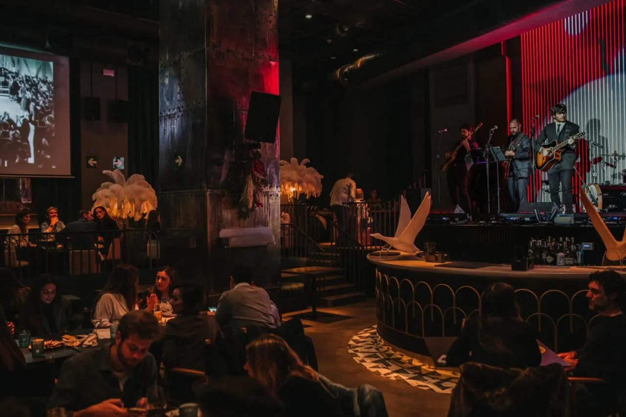 Restaurante con banda en vivo, decoración elegante, luces cálidas y pantalla con foto antigua. Ambiente animado y sofisticado.
