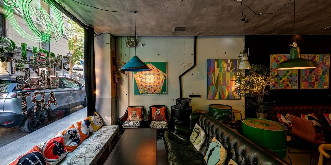 Interior de café moderno con arte geométrico, cojines coloridos, lámparas industriales y letrero “SMELLS LIKE ART” en ventana.