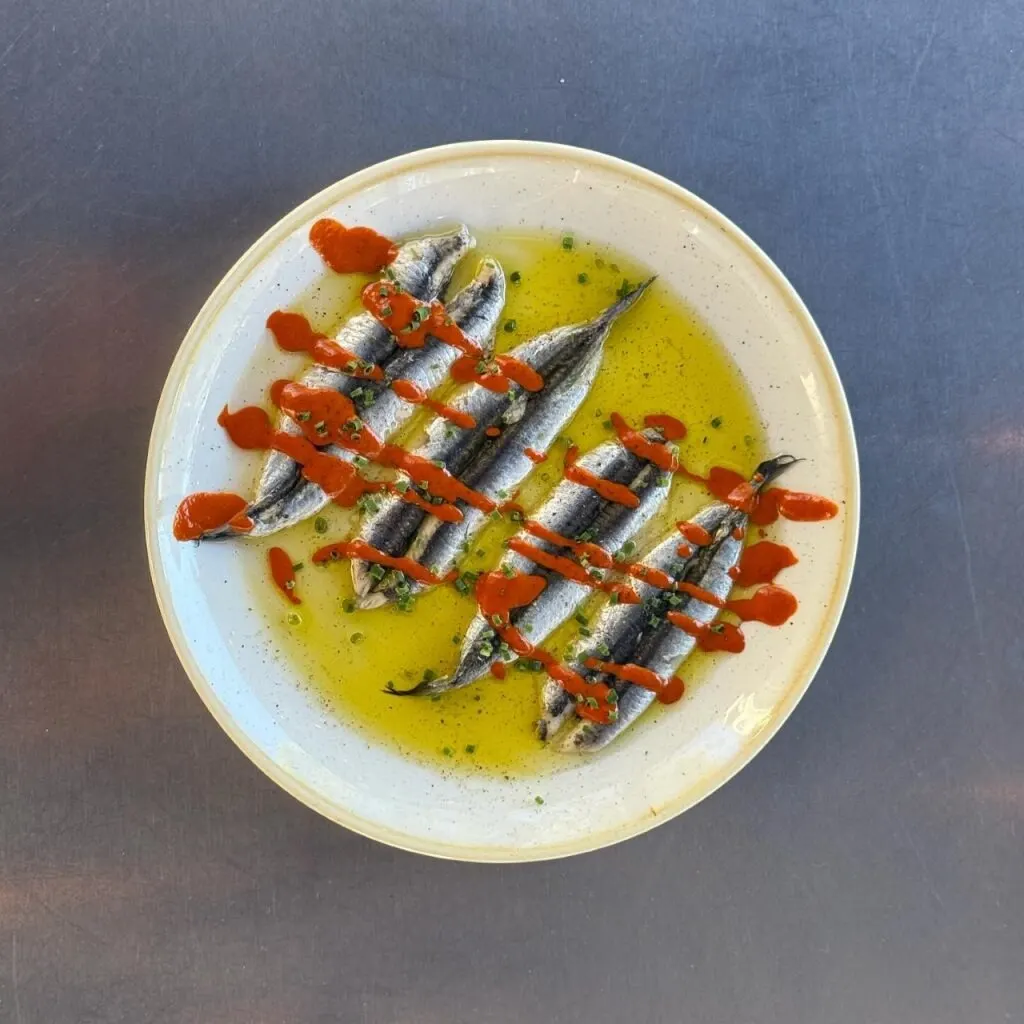 Sardinas en aceite dispuestas en círculo con salsa roja en cruz sobre plato blanco, apoyado en superficie metálica.