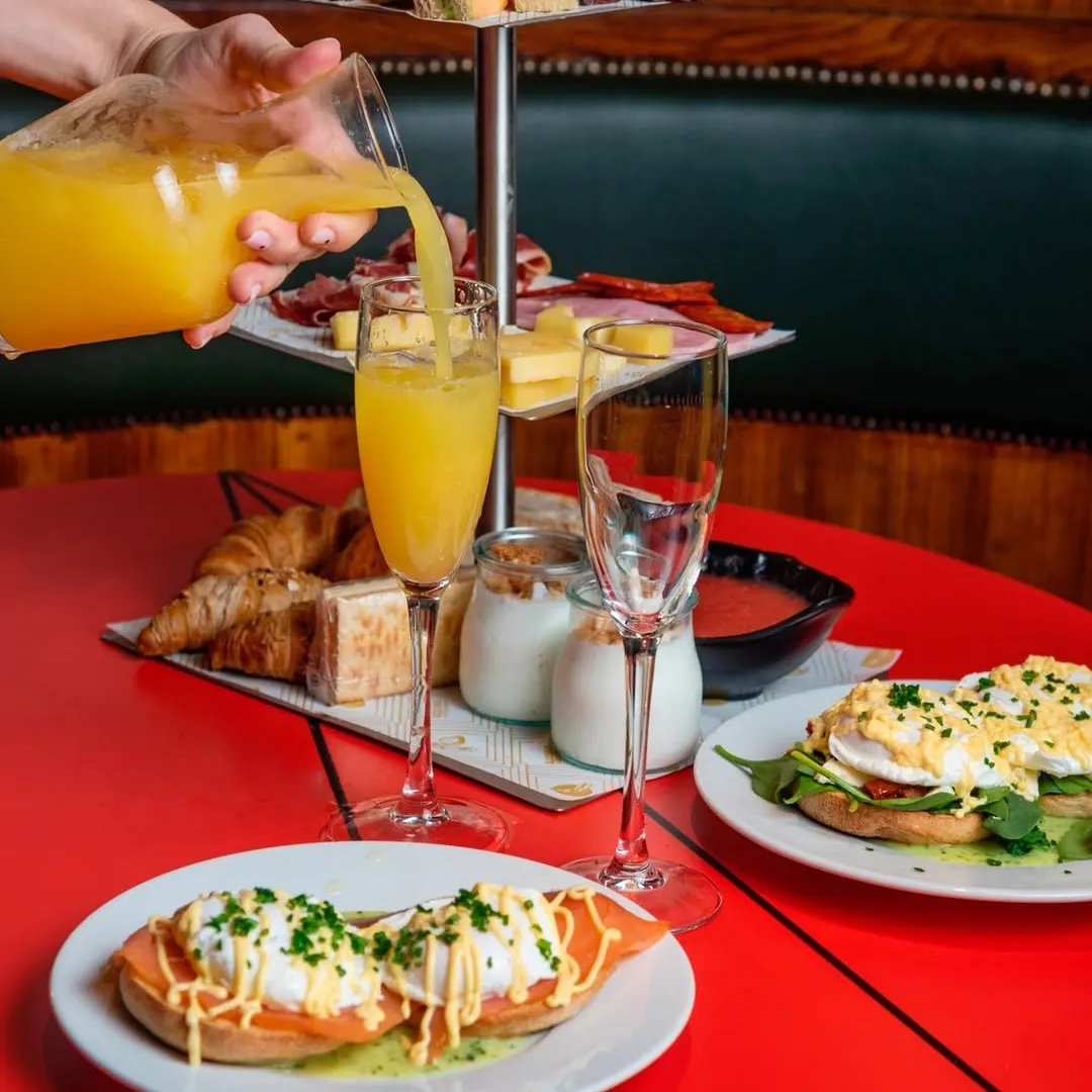 Mesa con desayuno completo, frutas, bollería, huevos benedictinos y zumo de naranja servido en copa junto a bandejas de embutido y yogur.