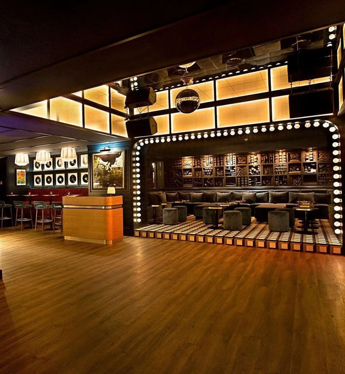 Bar elegante con suelo de madera, barra pequeña, sofás negros y luces redondas en arco. Decoración moderna con estanterías, cuadros y bola de discoteca.