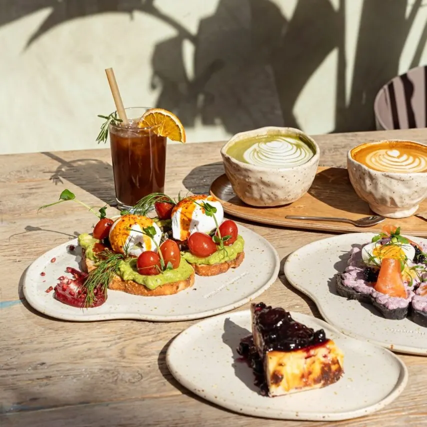 Mesa de madera con latte verde y naranja, bebida fría, tostadas con aguacate, salmón, cheesecake con frutos rojos y sombras de hojas.