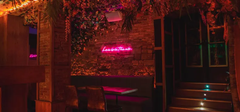 Mesa "booth" con sofá de terciopelo elegante, luces cálidas de ambiente y decoración sobria con paredes de piedra con cartel de neon. 