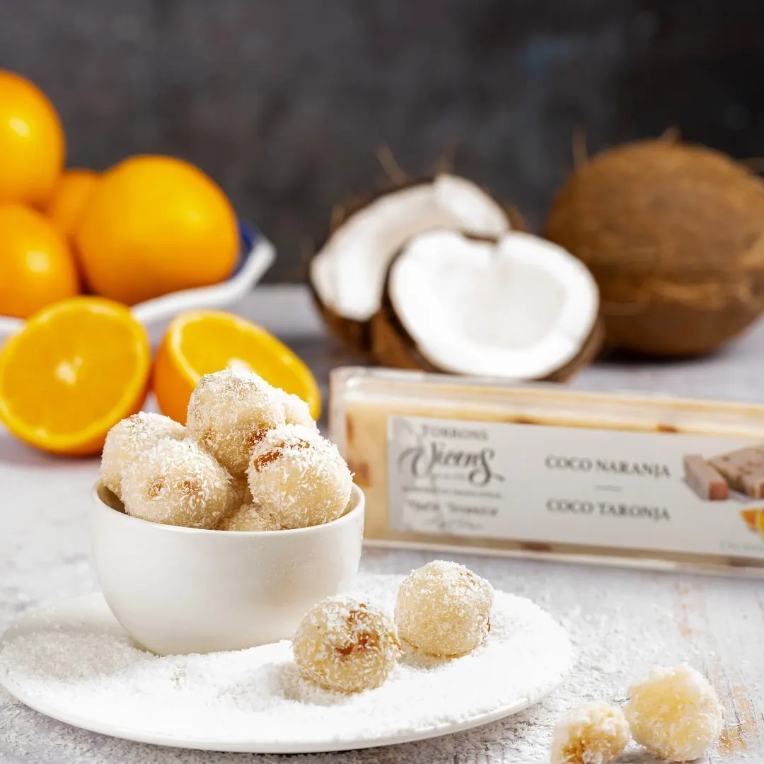 Bolas de coco cubiertas de ralladura fina servidas en un cuenco blanco, acompañadas de naranjas frescas y coco natural, junto a una barra de turrón de coco y naranja de Torrons Vicens.
