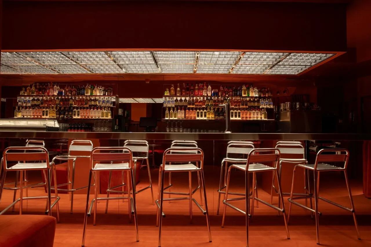 Barra iluminada de Gilda Haus con taburetes metálicos y estanterías llenas de botellas, todo rodeado de una estética rojiza y líneas geométricas.