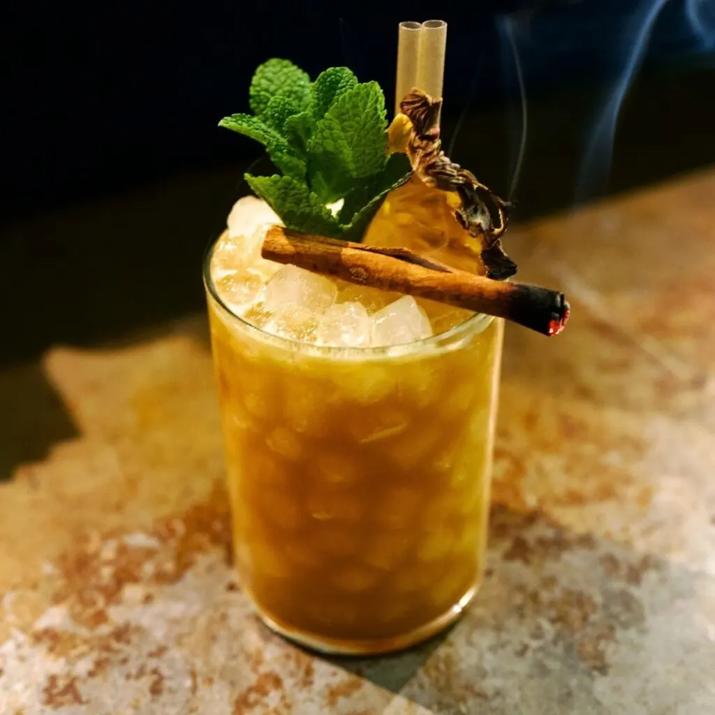 Cóctel con hielo, menta, canela y hierba humeante como decoración. Dos pajitas en vaso sobre superficie texturizada y fondo oscuro.
