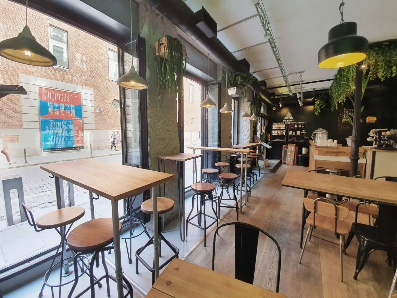 Interior de cafetería con mesas de madera, taburetes altos, plantas colgantes y grandes ventanales con vistas a la calle adoquinada.