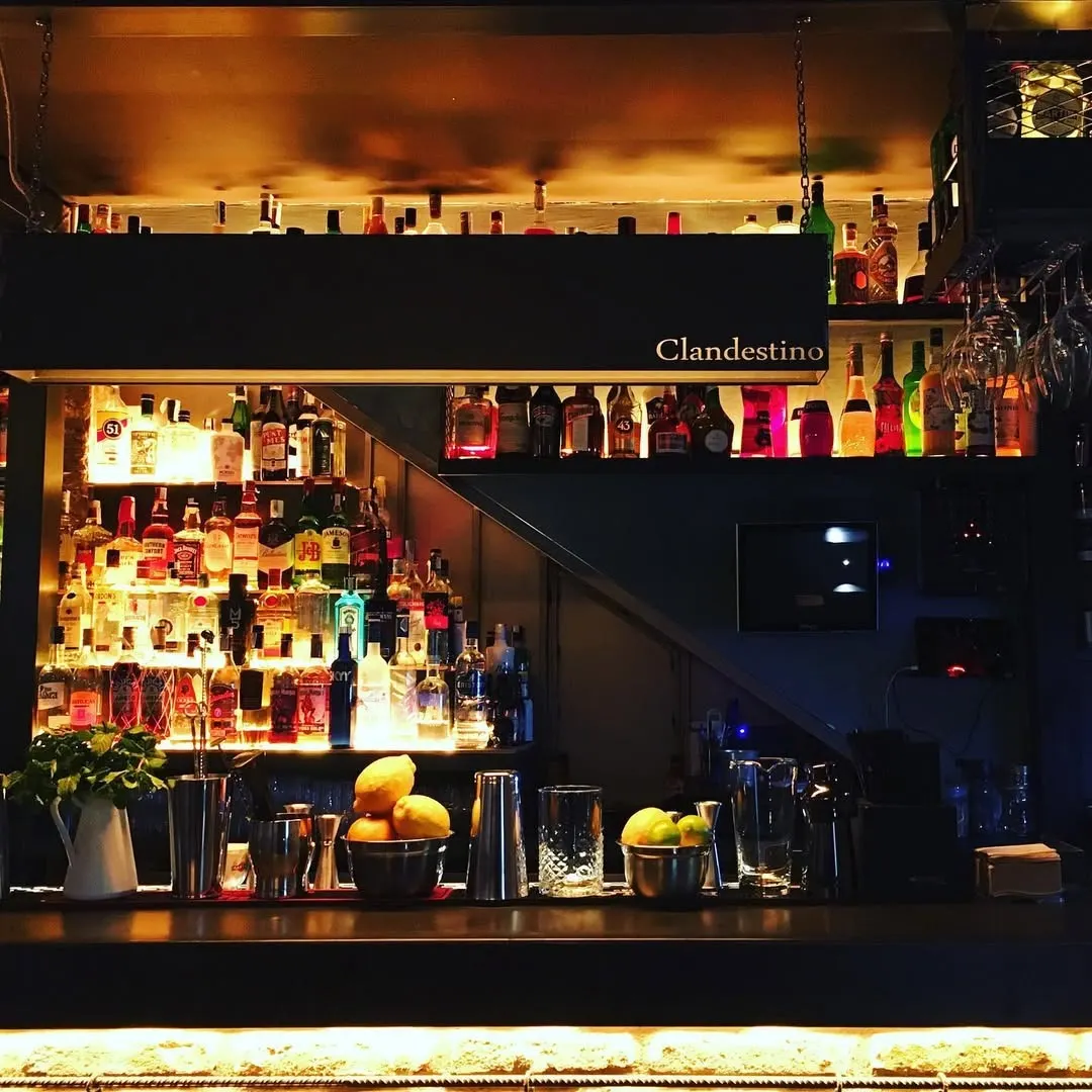 Botellas iluminadas y utensilios de coctelería en barra con luz cálida y cartel “Clandestino” en una atmósfera oscura y sofisticada.
