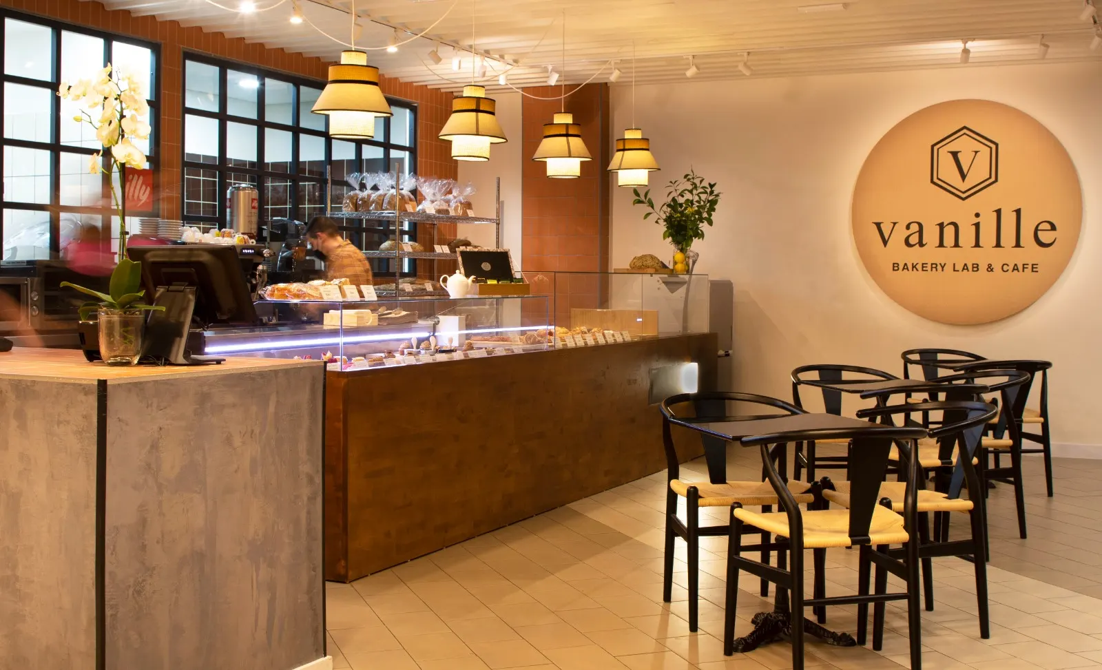 Interior de Vanille Bakery Lab con mostrador de madera lleno de repostería, lámparas colgantes cálidas y zona de mesas negras junto a un gran logo en la pared.