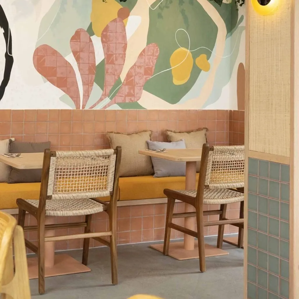 Zona de café con sillas de madera, banco con cojines, mural colorido, plantas colgantes y columna con azulejos teal y luz cálida.