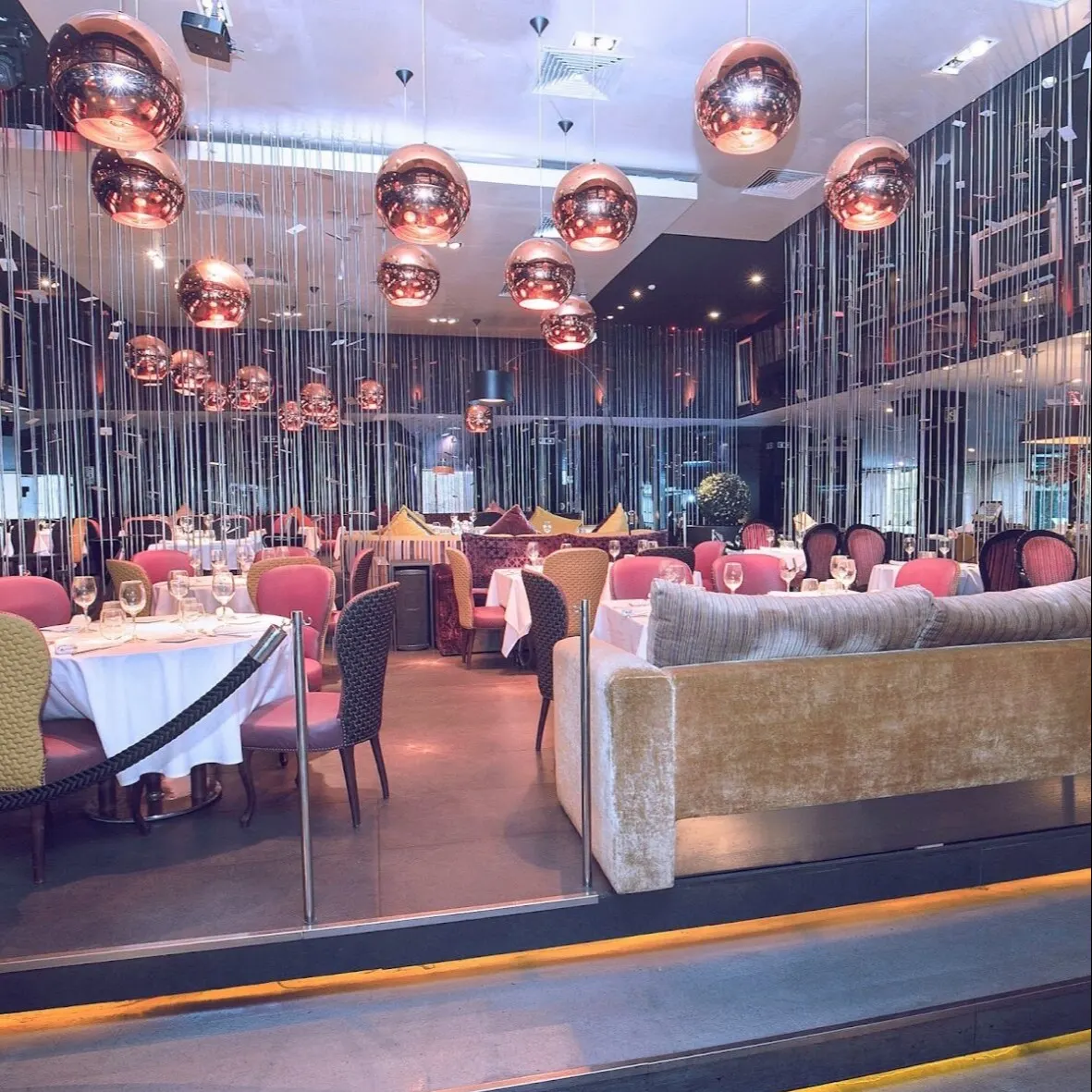 Restaurante moderno con lámparas metálicas, mesas con manteles blancos, copas, sillas rosas y paredes con espejos.