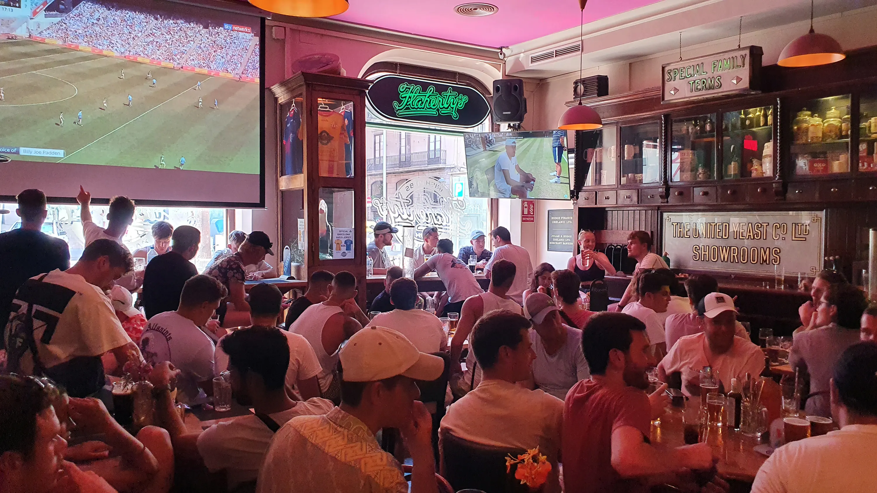 La imagen muestra el interior del flaherty´s. Hay una larga barra roja con varios taburetes frente a ella. Detrás de la barra, hay estantes llenos de numerosas botellas de alcohol que llegan hasta el techo. 