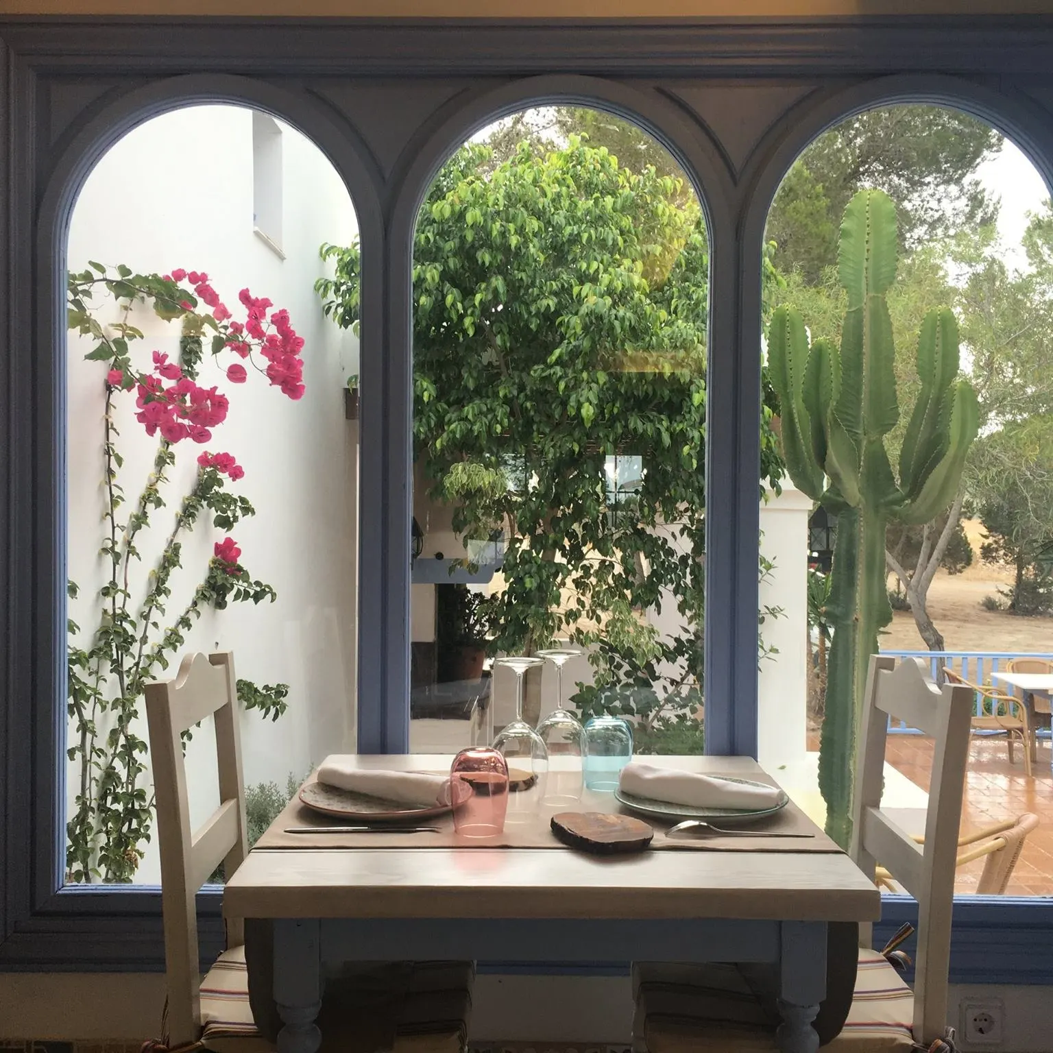 La imagen muestra la tranquila atmósfera de Es Tragon, con una mesa preparada junto a una ventana que enmarca un jardín lleno de flores y vegetación
