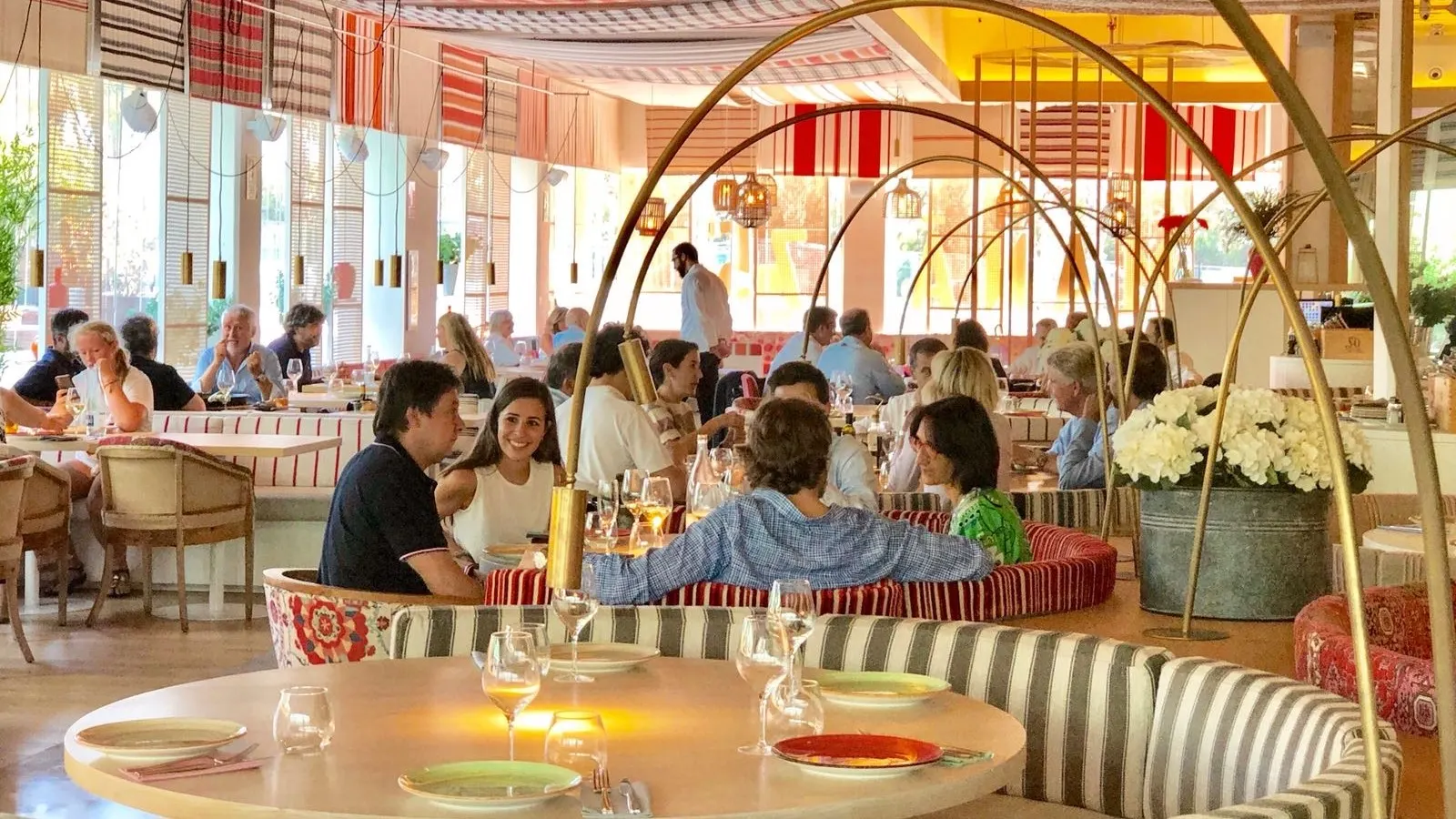 La imagen muestra un área de comedor del Maná 75, con una decoración vibrante y colorida. Hay varias mesas preparadas con platos, vasos y cubiertos, listas para los comensales. El techo tiene patrones de rayas en rojo y blanco.