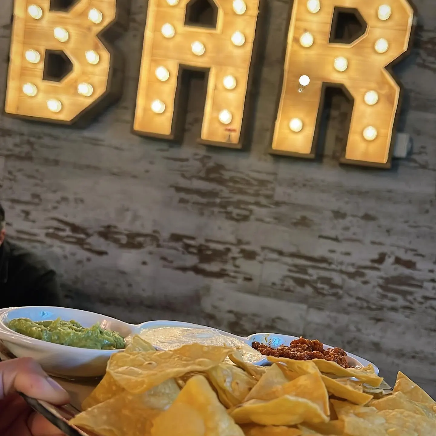 Plato de nachos con guacamole, crema y carne, sostenido frente a un cartel iluminado con letras que forman la palabra "BAR".