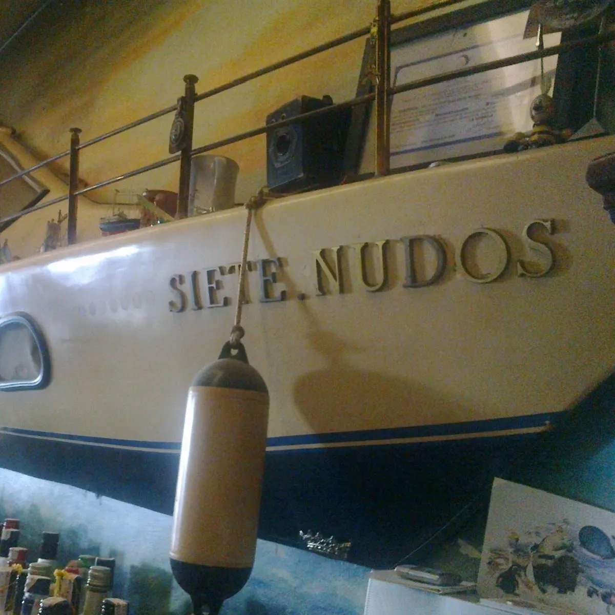 Barra con botellas frente a una gran maqueta de barco en la pared, con el nombre "Siete Nudos" y detalles náuticos decorativos.