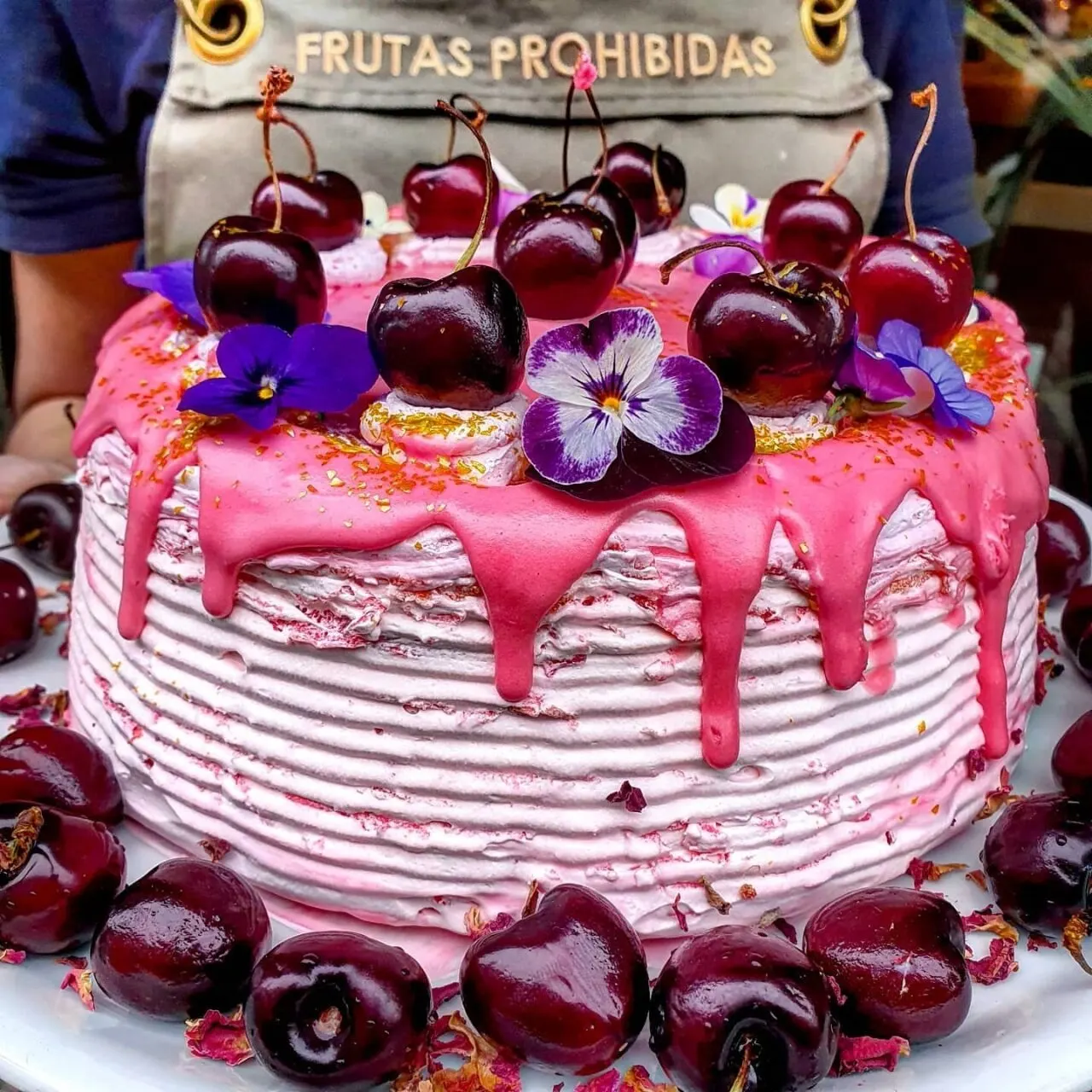 Tarta con glaseado rosa y cerezas, decorada con flores moradas. Está sobre plato blanco, rodeada de cerezas. Fondo con delantal “FRUTAS PROHIBIDAS”.