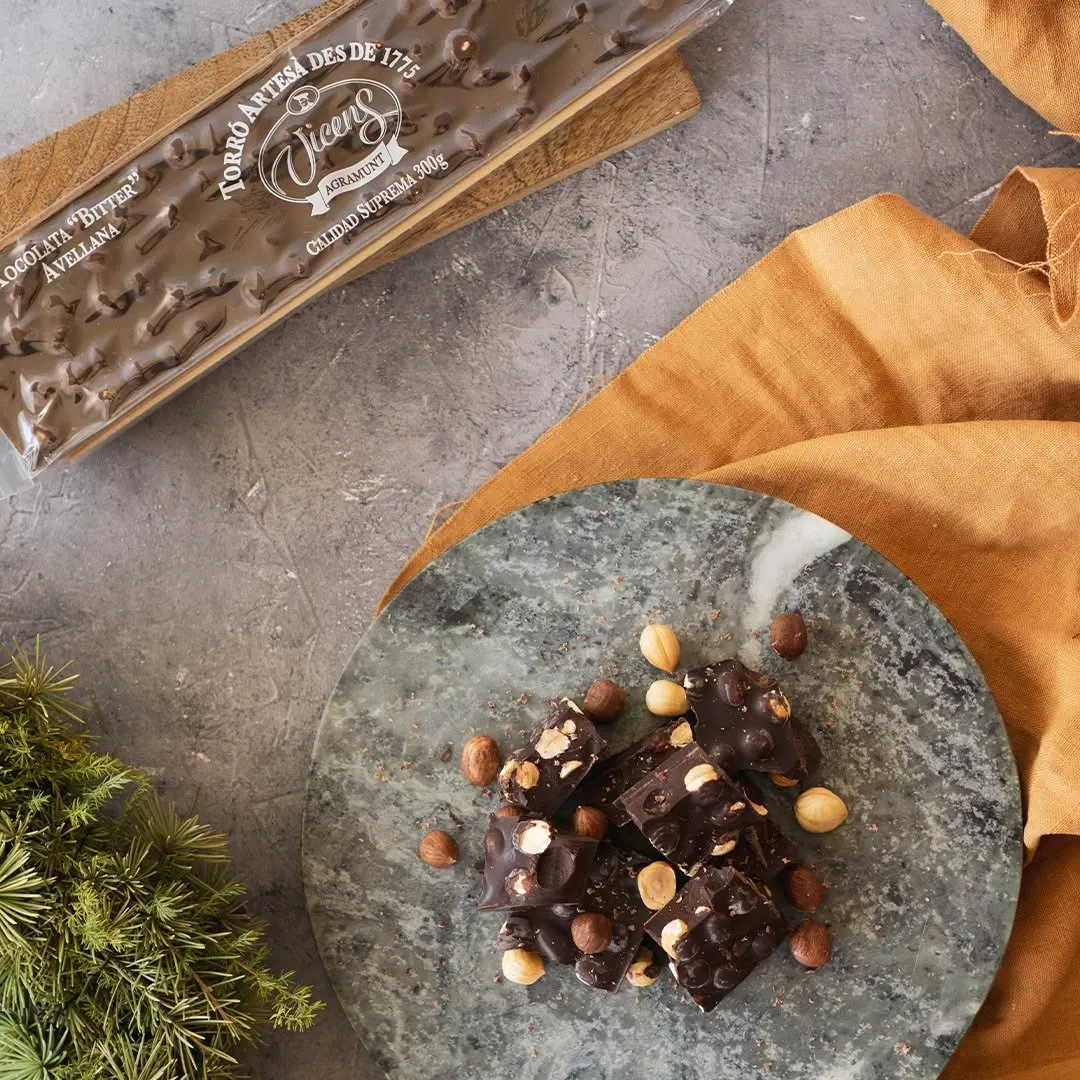 Trozos de turrón de chocolate bitter con avellanas sobre un plato de mármol verde, junto a su tableta original de Torrons Vicens envuelta y una base decorativa de tela mostaza.