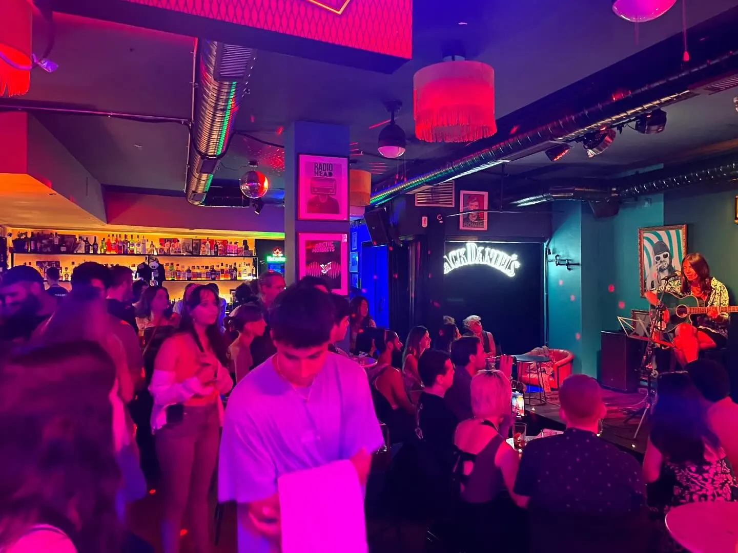 La imagen muestra un bar animado con luces de colores vibrantes. En el escenario, un músico toca la guitarra ante un público atento. Al fondo, se observan botellas en el estante del bar, creando una atmósfera acogedora y festiva.