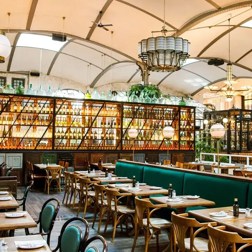 Amplio salón en El Nacional Barcelona, con mesas de madera, sofás verdes, botellas exhibidas y una decoración elegante con plantas colgantes.