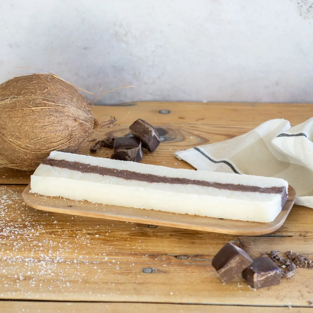 Turrón de coco con capa central de chocolate sobre bandeja de madera, acompañado de virutas, trozos de chocolate y un coco entero sobre fondo rústico y natural.