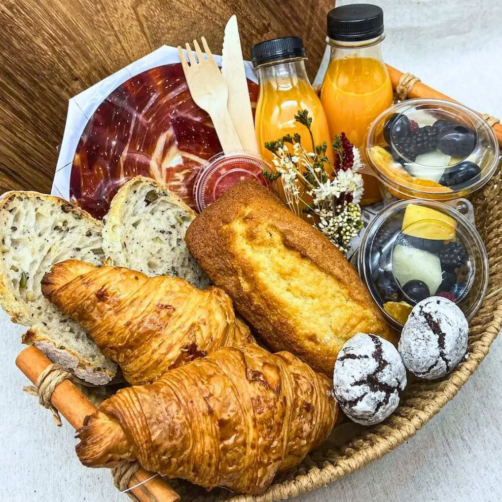 Cesta de desayuno con croissants, bizcocho, pan de semillas, fruta fresca, zumo de naranja, dulces y jamón en lonchas sobre fondo de madera.