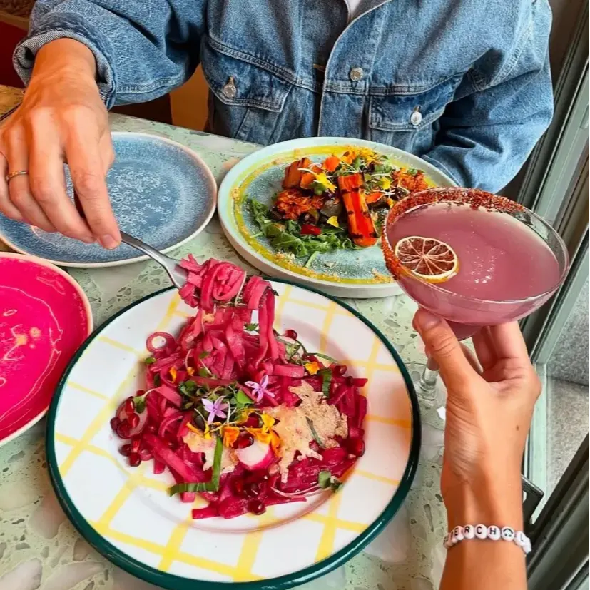 Dos personas cenan juntas; una sostiene un cóctel rosa con limón seco, la otra lleva chaqueta vaquera. Hay pasta rosa y verduras a la parrilla.