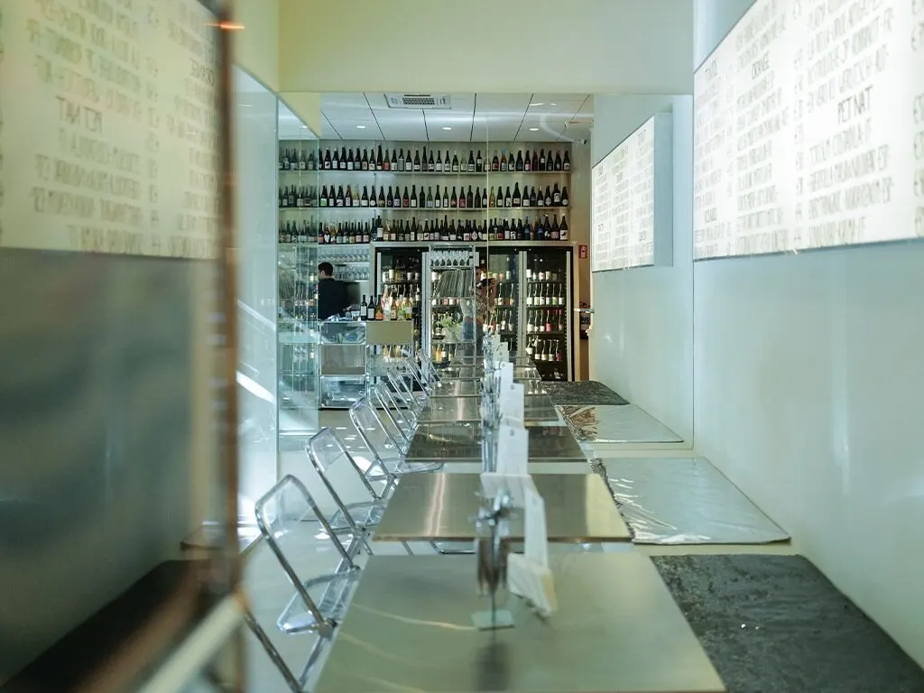 Interior de Masa Madrid con mesas de acero alineadas, sillas metálicas y estanterías repletas de botellas que dominan el fondo del espacio con un estilo minimalista.